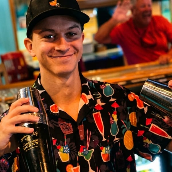 Smiling bartender in a colorful shirt holding two cocktail shakers, with customers in the background.
