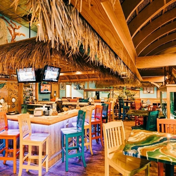 Tropical-themed restaurant interior with a thatched bar, colorful chairs, and mermaid mural on a blue wall.